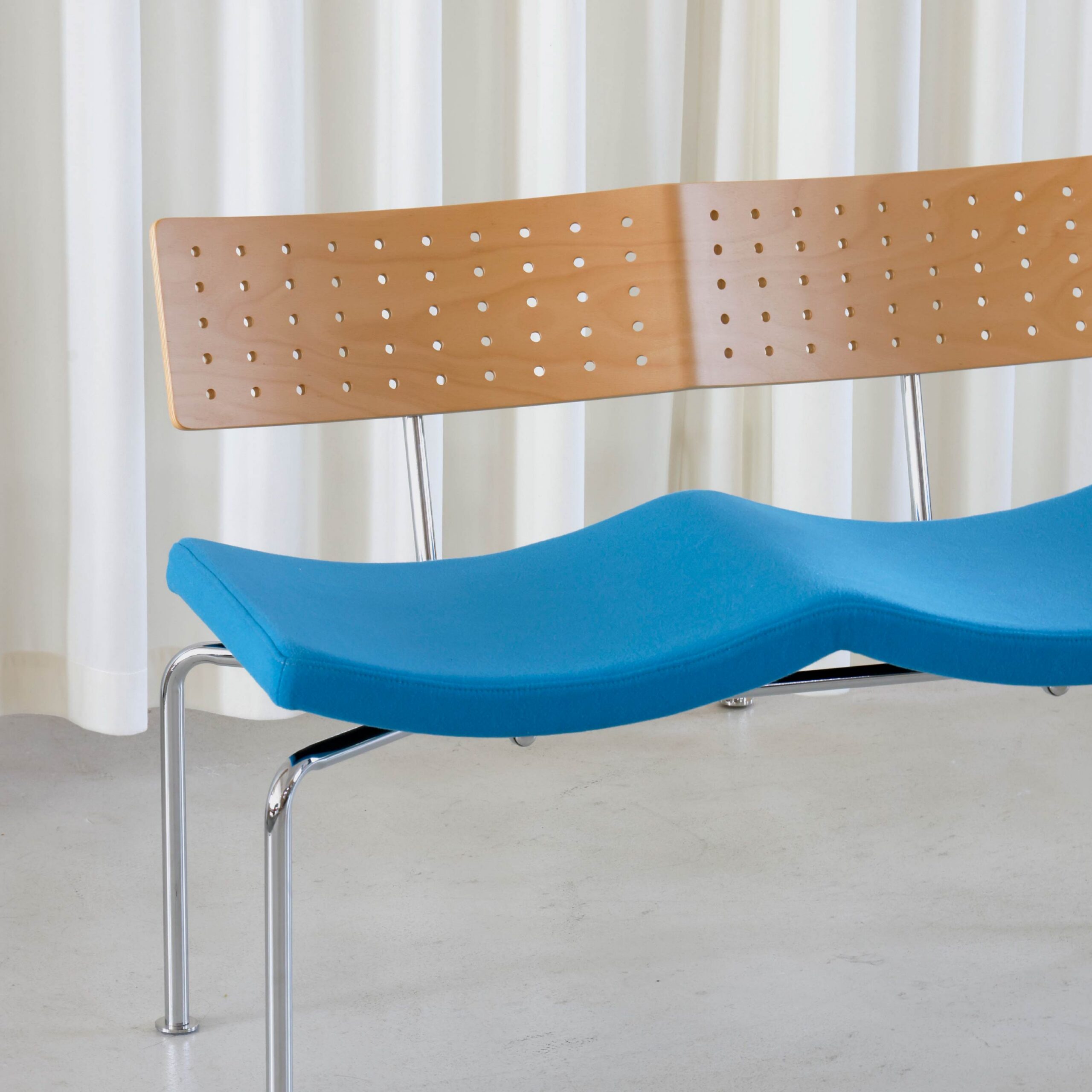 A modern bench with a contoured blue seat, a perforated wooden backrest, and metal legs, placed on a light floor in front of white curtains.