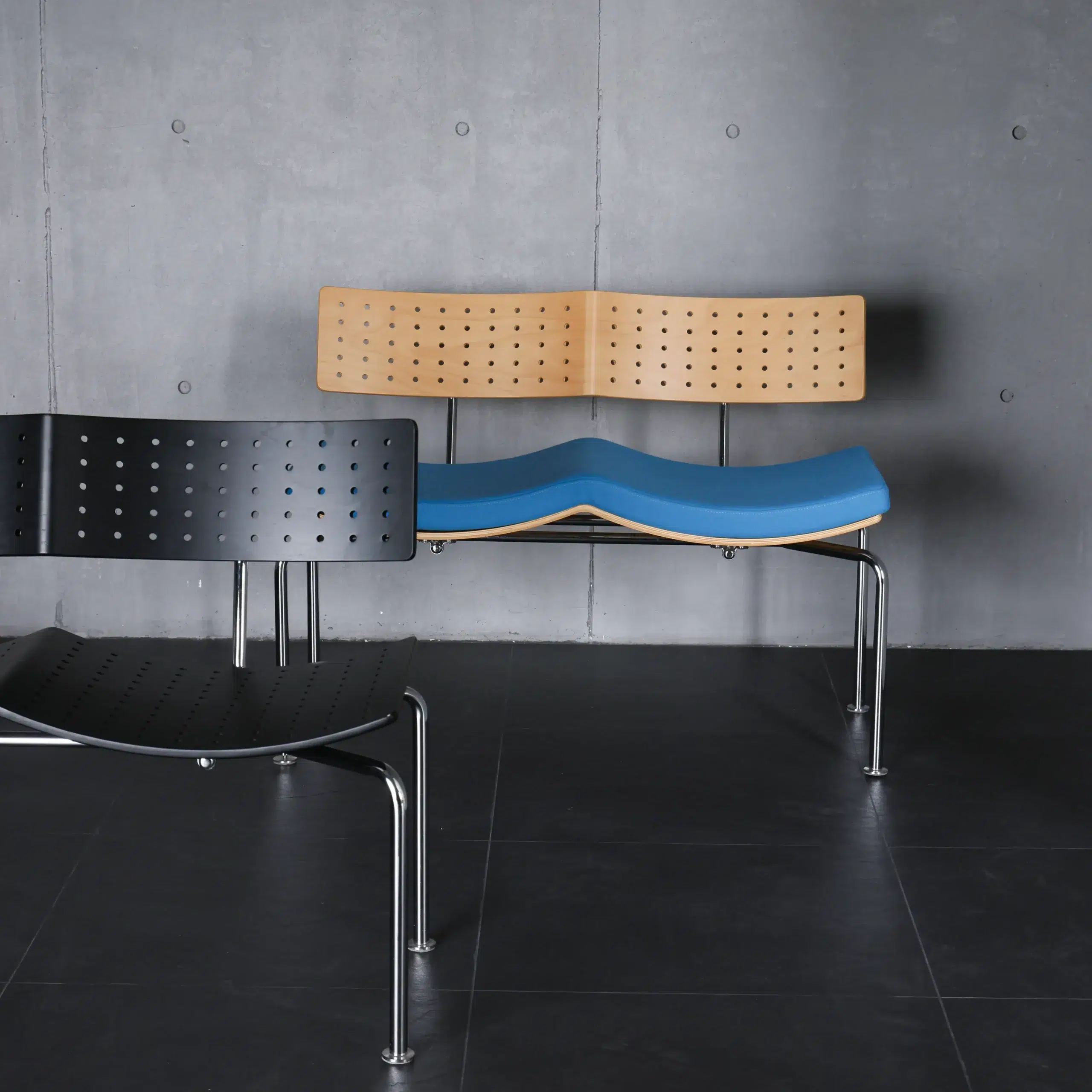 Two modern chairs with perforated backs and metal legs sit on a dark tiled floor. One chair is black, and the other has a light wood back with a blue curved seat. The background is a grey concrete wall.
