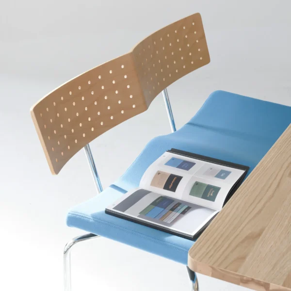 A modern chair with a blue seat and perforated wooden backrest sits next to a light wood table. An open magazine with colourful pages rests on the chair's seat.
