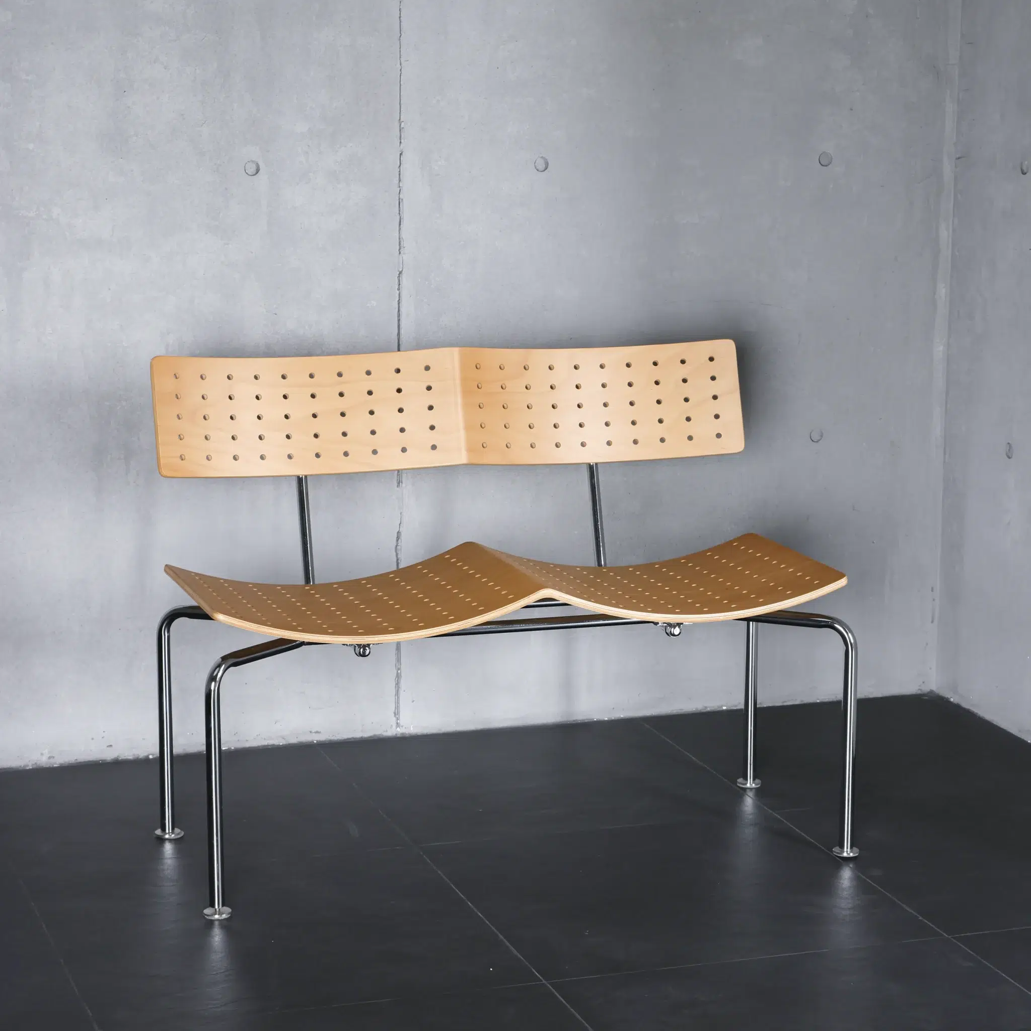 A modern wooden bench with a curved seat and backrest featuring small holes, supported by slim metal legs, is placed against a grey concrete wall on a dark tiled floor.
