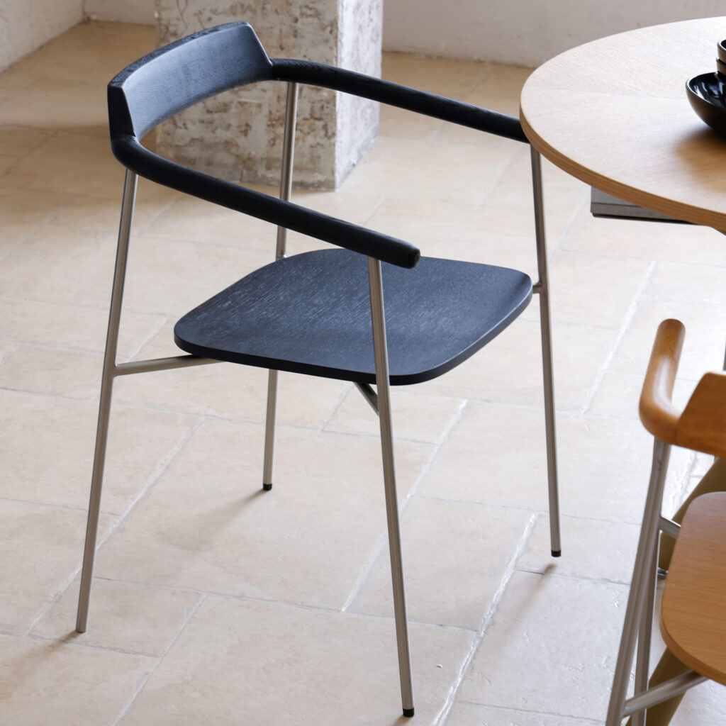 A modern chair with a black seat and armrests, featuring thin, light-coloured metal legs. The chair is placed on a tiled floor next to a round wooden table and another light wood chair.