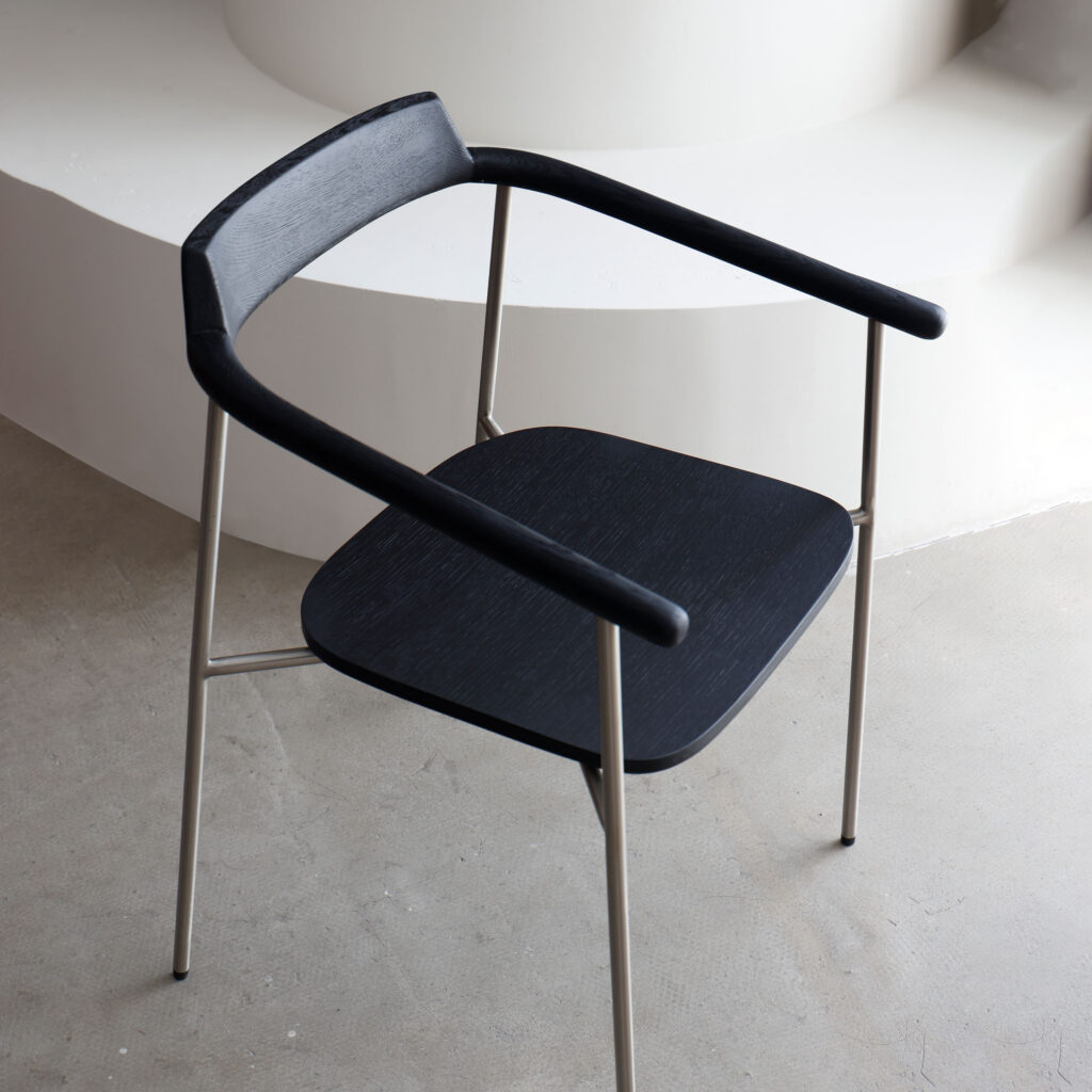 A modern chair with a black wooden seat and backrest, featuring thin, metallic legs and armrests. The chair is placed on a light-coloured floor near curved, white architectural steps.