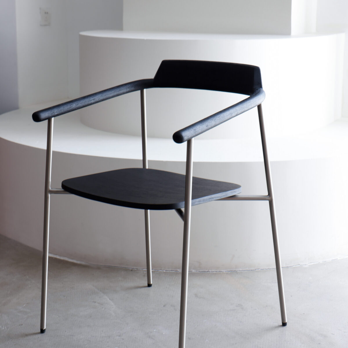 Gentleman Dining Chair_Black_C6 A modern black chair with a minimalist design, featuring a wooden seat and backrest, thin metal legs, and armrests, positioned in front of white curved steps in a bright room.