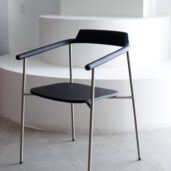 A modern black chair with a minimalist design, featuring a wooden seat and backrest, thin metal legs, and armrests, positioned in front of white curved steps in a bright room.