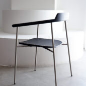 A modern minimalist chair with a black wooden seat and backrest, slim metal legs, and an open angular design, placed on a grey floor in front of a white curved staircase.