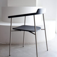 A modern minimalist chair with a black wooden seat and backrest, slim metal legs, and an open angular design, placed on a grey floor in front of a white curved staircase.