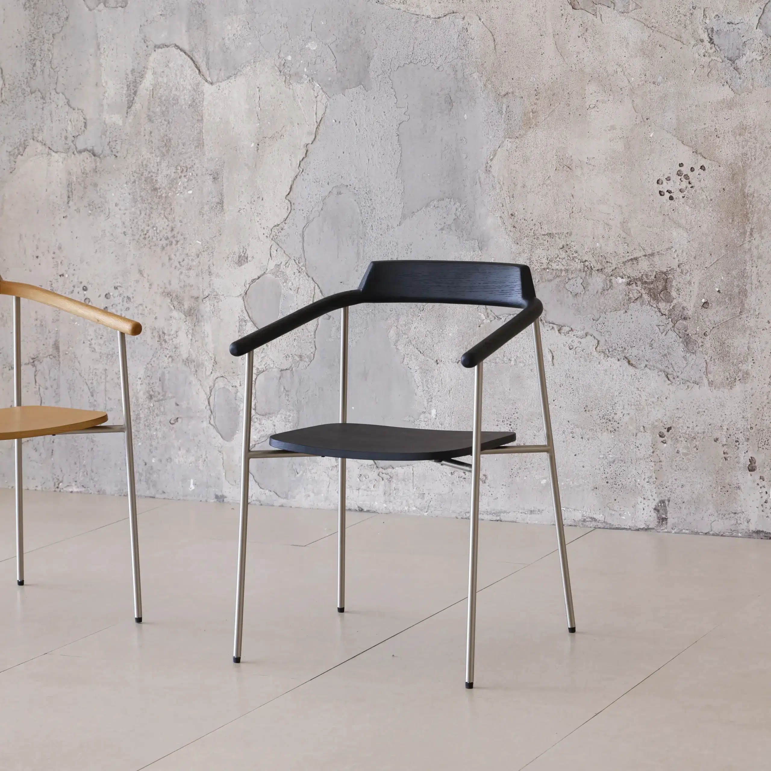 A minimalist black chair with thin metal legs and armrests is placed on a light tiled floor next to a partial view of a similar tan chair, against a textured, cracked concrete wall.