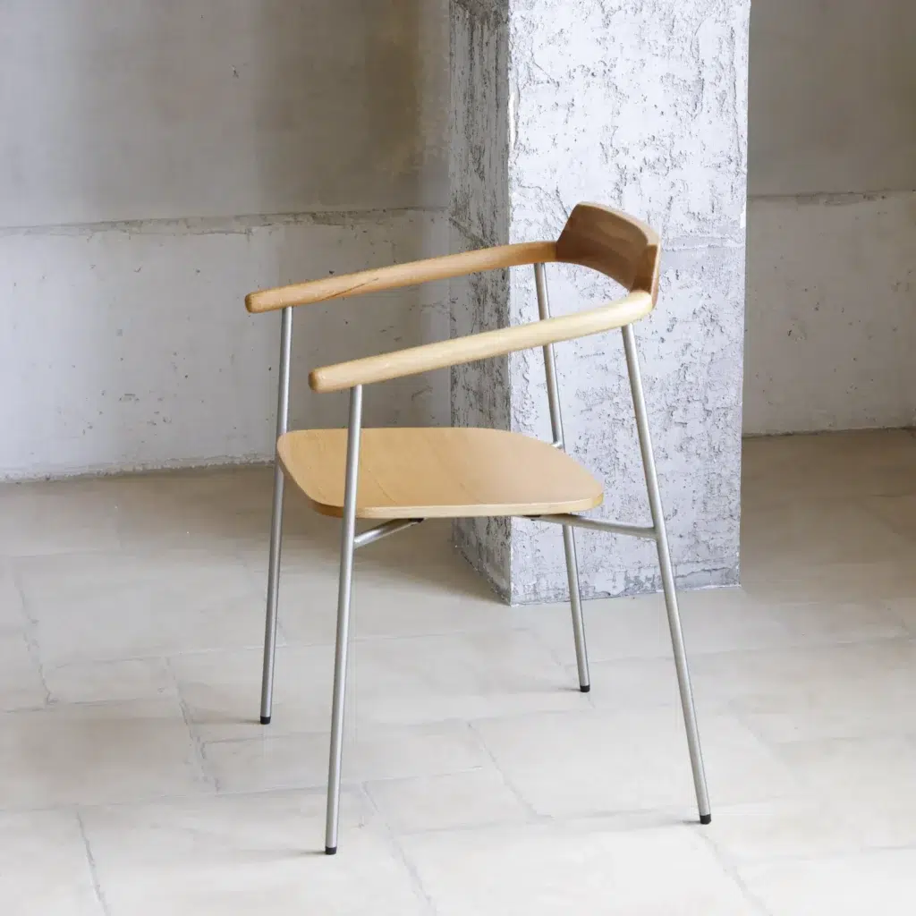 A minimalist wooden chair with a curved backrest and metal legs stands on a light tiled floor against a textured white wall and pillar.
