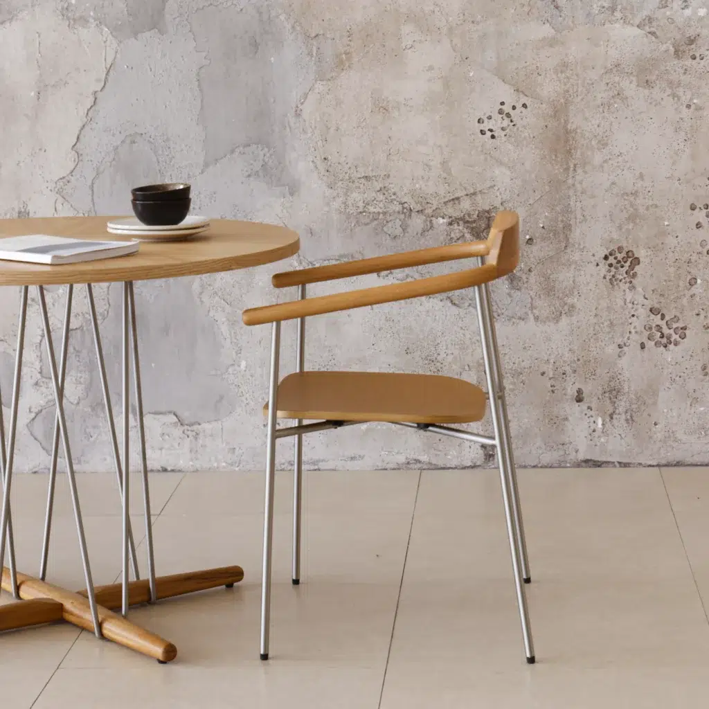 A wooden chair with metal legs is placed next to a round wooden table with thin metal supports. On the table are a stacked cup and saucer, a closed notebook, and a pen. The background features a textured concrete wall.