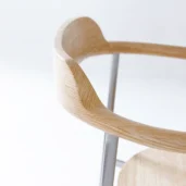 Close-up of a modern wooden chair with a curved, light-coloured wooden backrest and metal supports, set against a plain white background.