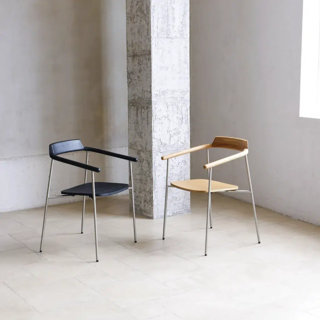 Two minimalist chairs with metal legs, wooden armrests, and angled backs—one with a dark seat and one with a light seat—are positioned near a concrete column in a bright, modern room.