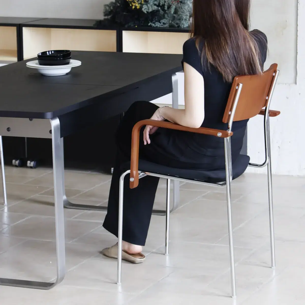 Knight Dining Chair_Back Side View(4) A woman with long brown hair, dressed in black, sits sideways on a chair at a black table with dishes on it in a modern, minimalist room with light tiled flooring.