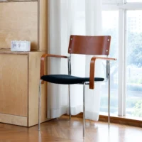 A modern chair with a brown wooden backrest, curved wooden armrests, black cushioned seat, and chrome legs is placed by a window with white curtains on a wooden floor near a light wood cupboard.