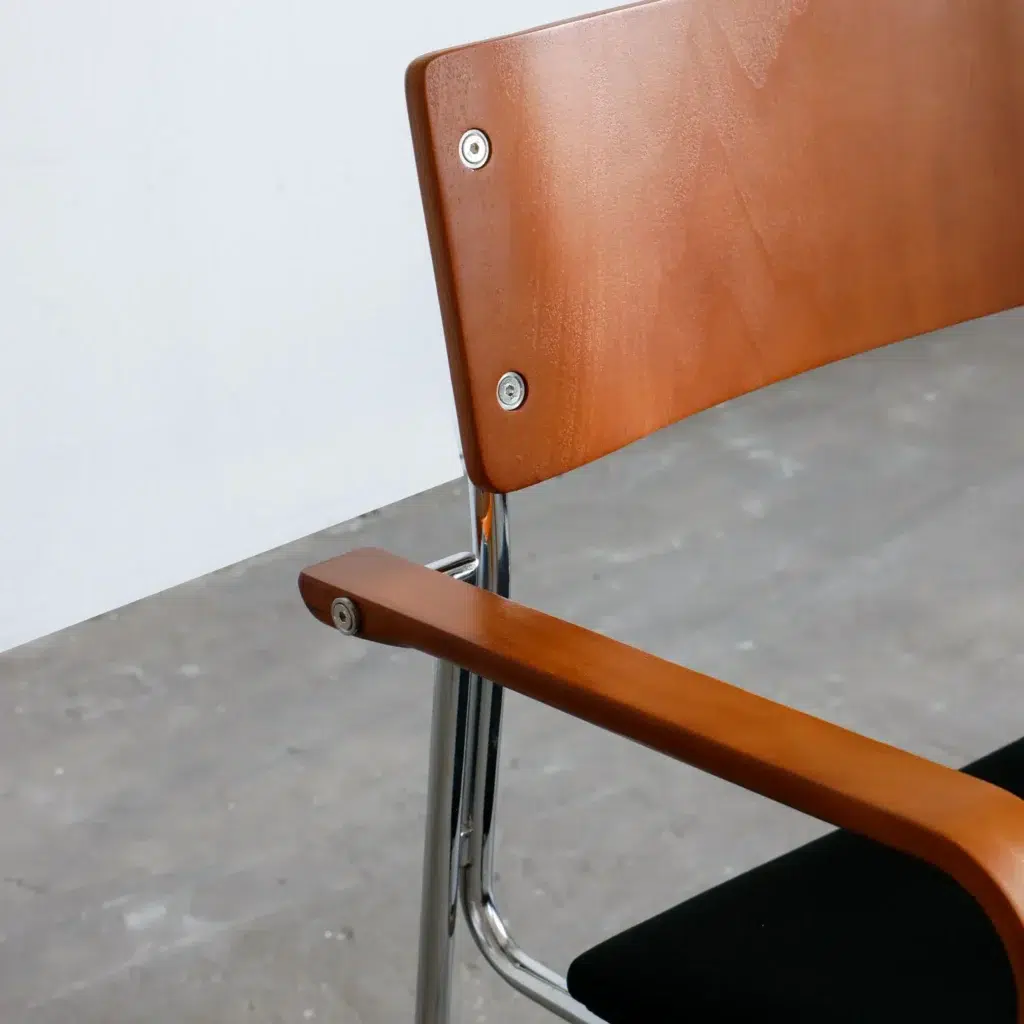 Close-up of a modern chair with a wooden backrest and armrest, chrome metal frame, and black cushioned seat, set on a grey concrete floor against a plain white wall.