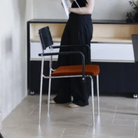A person in black trousers and a black top stands by a modern chair with a red seat and metal legs, holding papers in an office setting with light flooring and a black-and-white cupboard.