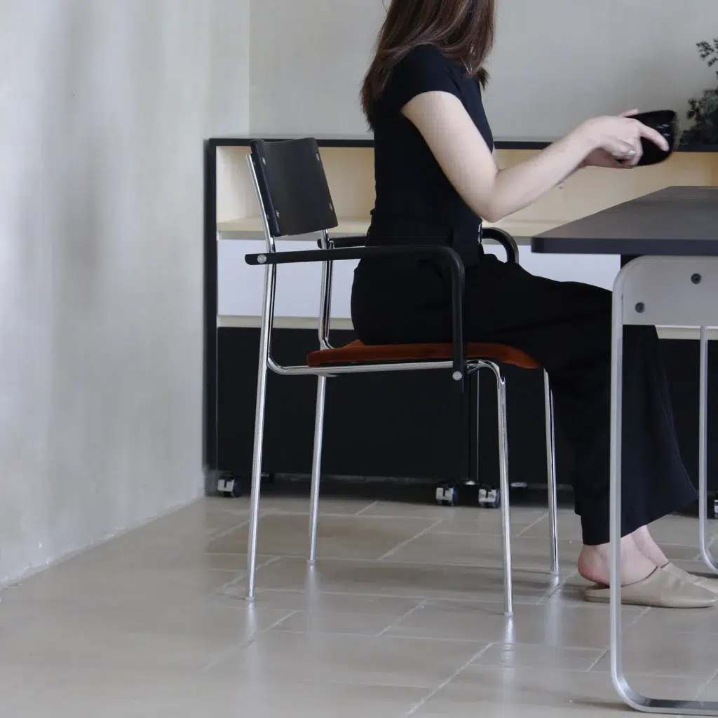 A woman with long hair, dressed in black, sits sideways on a chair at a table, holding an object in her hand. The room has light-coloured walls and floor tiles. Her face is not visible.
