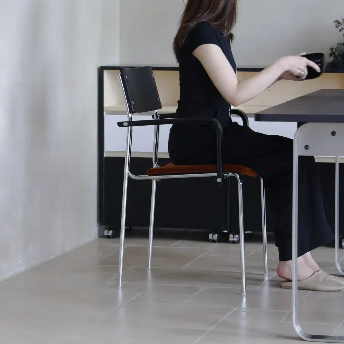 Knight Dining Chair_Side View(2) A woman with long hair, dressed in black, sits sideways on a chair at a table, holding an object in her hand. The room has light-coloured walls and floor tiles. Her face is not visible.