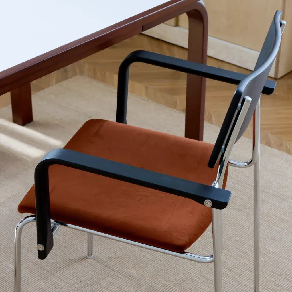 A modern chair with a brown seat cushion, black armrests, and metal legs sits beside a wooden table on a beige rug with a parquet floor in the background.