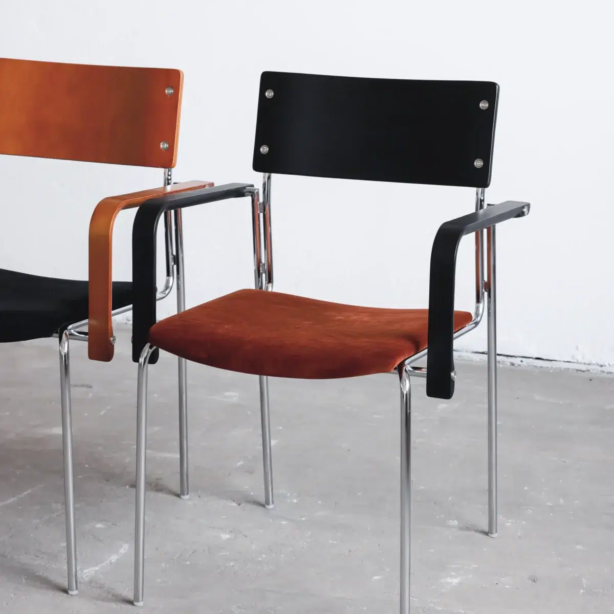 Knight Dining Chair__Black_Front Side View Two modern chairs with metal legs; one has a black backrest and brown seat, the other has a brown backrest and black seat. Both have black armrests and are placed on a grey concrete floor against a white wall.