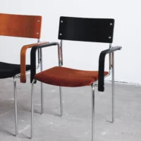 Two modern chairs with metal legs; one has a black backrest and brown seat, the other has a brown backrest and black seat. Both have black armrests and are placed on a grey concrete floor against a white wall.