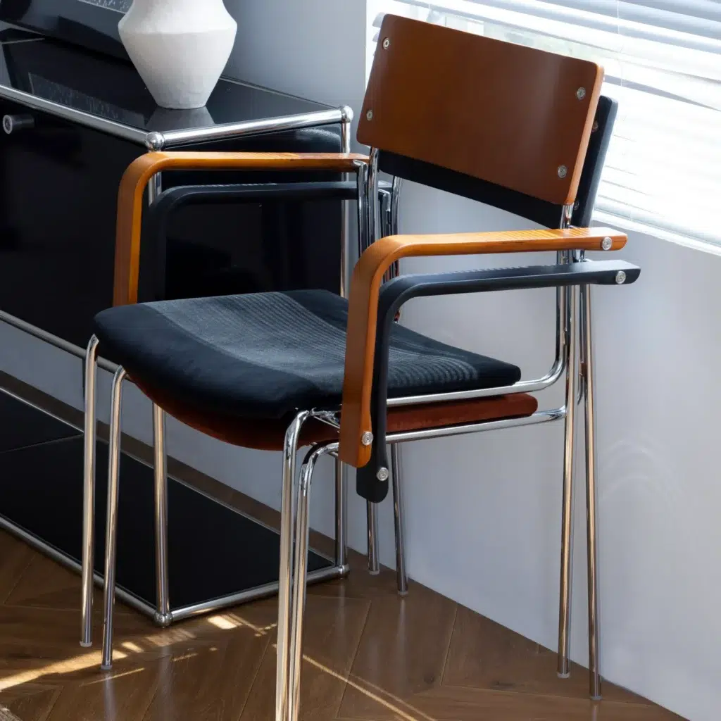A modern chair with a wooden backrest, black seat cushion, and chrome legs is positioned near a window with closed blinds. The chair is next to a black cupboard on a wooden floor.