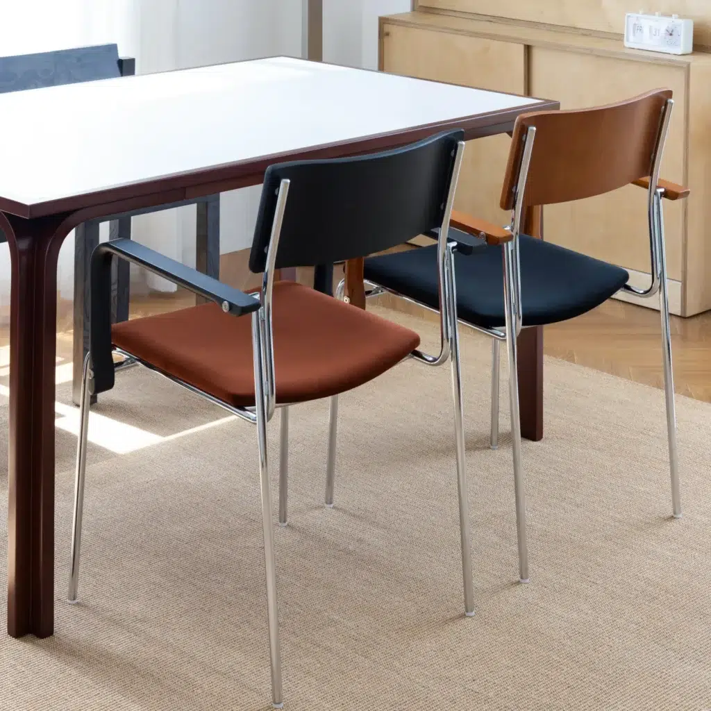 Two modern chairs with chrome legs and coloured seats—one brown and one black—are placed side by side at a white rectangular table in a bright, minimalist room with wooden flooring and a beige rug.