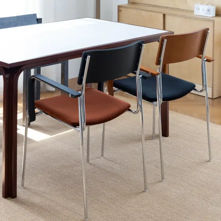 Two modern chairs with chrome legs and coloured seats—one brown and one black—are placed side by side at a white rectangular table in a bright, minimalist room with wooden flooring and a beige rug.