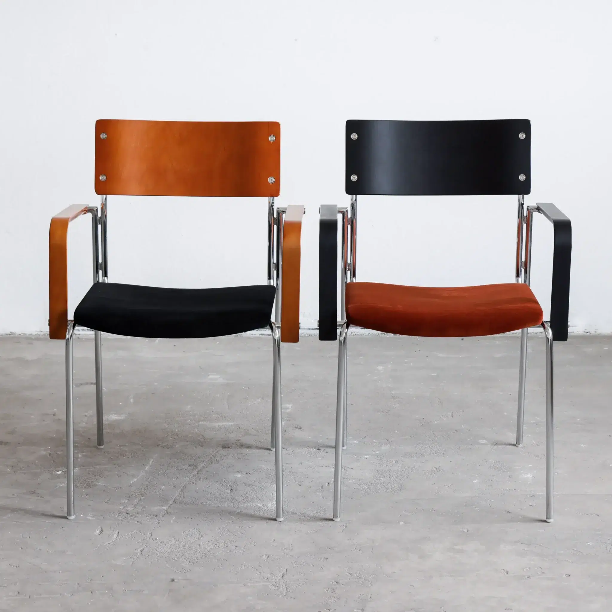 Two modern chairs with metal legs and armrests. The left chair has a brown wooden backrest and armrests with a black seat, whilst the right chair has a black backrest and armrests with a brown seat. They are on a grey floor.