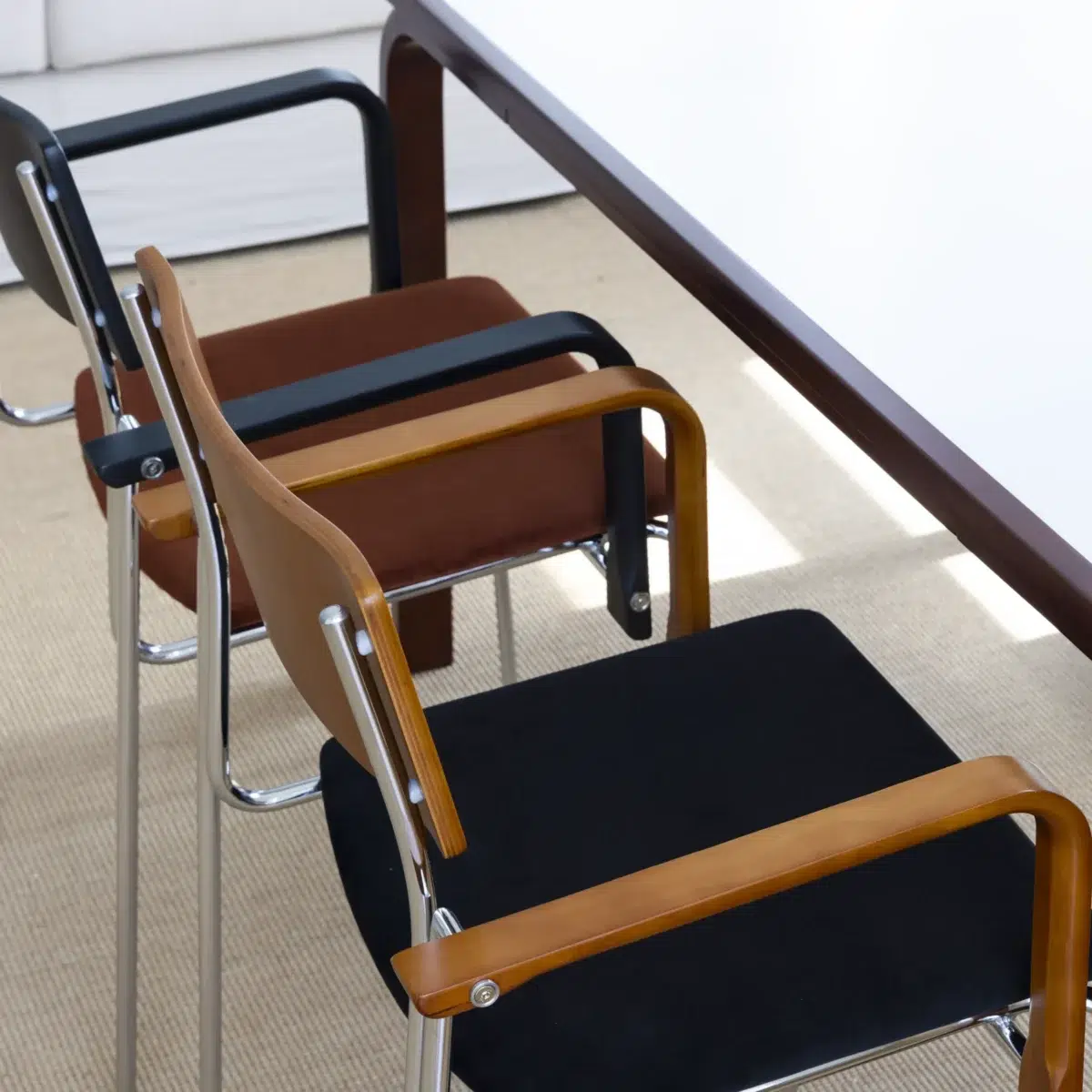 Knight Dining Chair__Pair_Side View Three modern chairs with wooden armrests and metal frames are placed beside a rectangular table on a light-coloured carpet. The seats and backs of the chairs are upholstered in black and brown fabric.