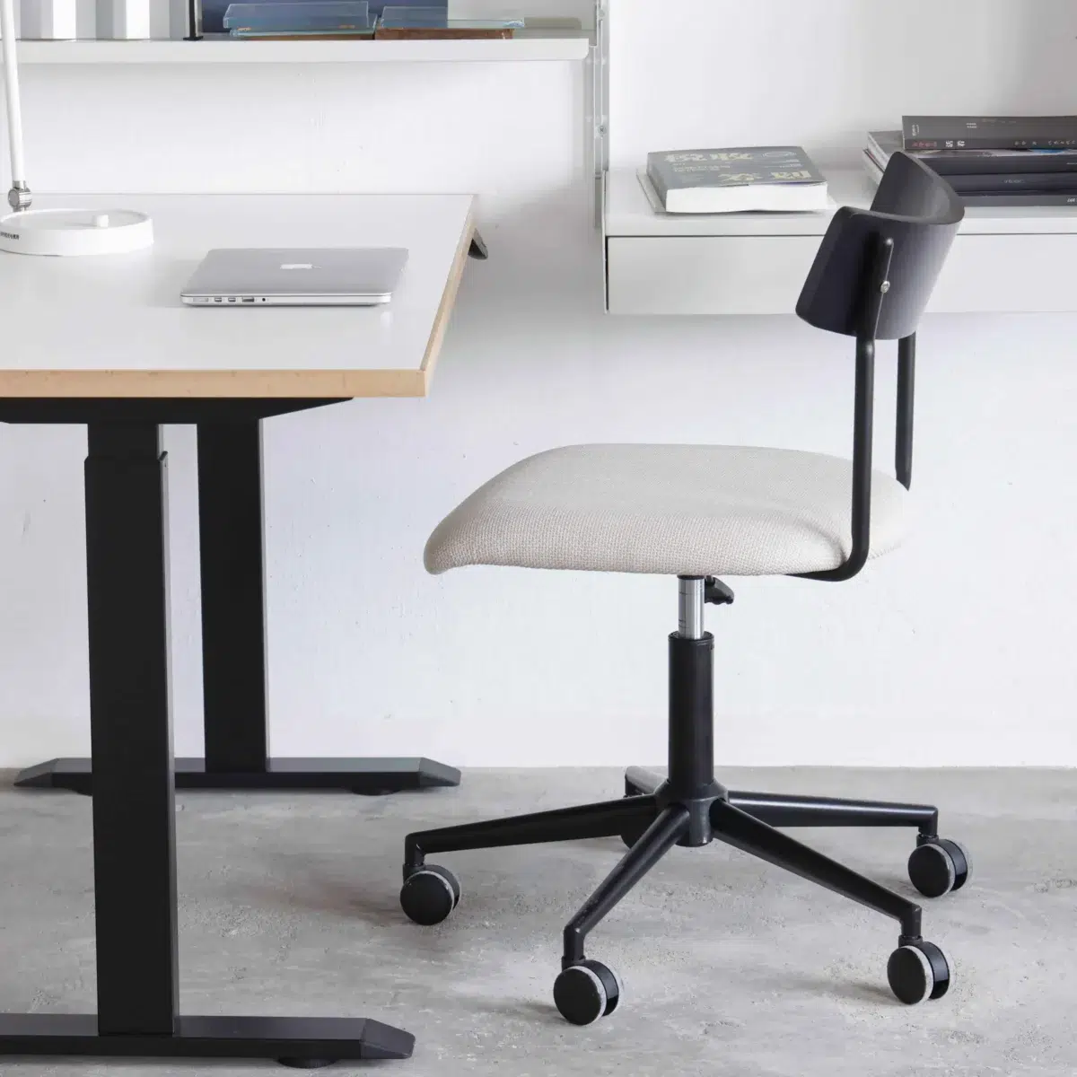 Lift Office Chair_Side View(4) A modern office set-up with a white desk, a closed laptop, and a rolling office chair with a black frame and light-coloured seat on a concrete floor. Shelves and books are visible in the background.