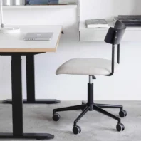 A modern office set-up with a white desk, a closed laptop, and a rolling office chair with a black frame and light-coloured seat on a concrete floor. Shelves and books are visible in the background.