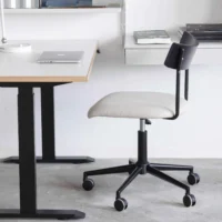A modern office set-up with a white desk, a closed laptop, and a rolling office chair with a black frame and light-coloured seat on a concrete floor. Shelves and books are visible in the background.