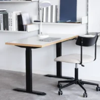 A modern desk with a laptop, white lamp, and office chair on castors. Minimalist shelves above hold books and folders. The setting is clean, organised, and well-lit, with a concrete floor and white wall.