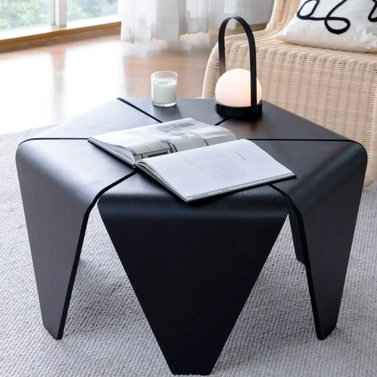 Lotus Coffee Table_Black A modern black coffee table with an open book, a lit candle, and a small round lamp on top, set on a light-coloured rug in a bright living room with a wicker chair and large windows in the background.