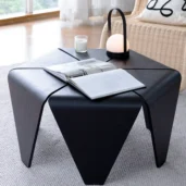 A modern black coffee table with an open book, a lit candle, and a small round lamp on top, set on a light-coloured rug in a bright living room with a wicker chair and large windows in the background.