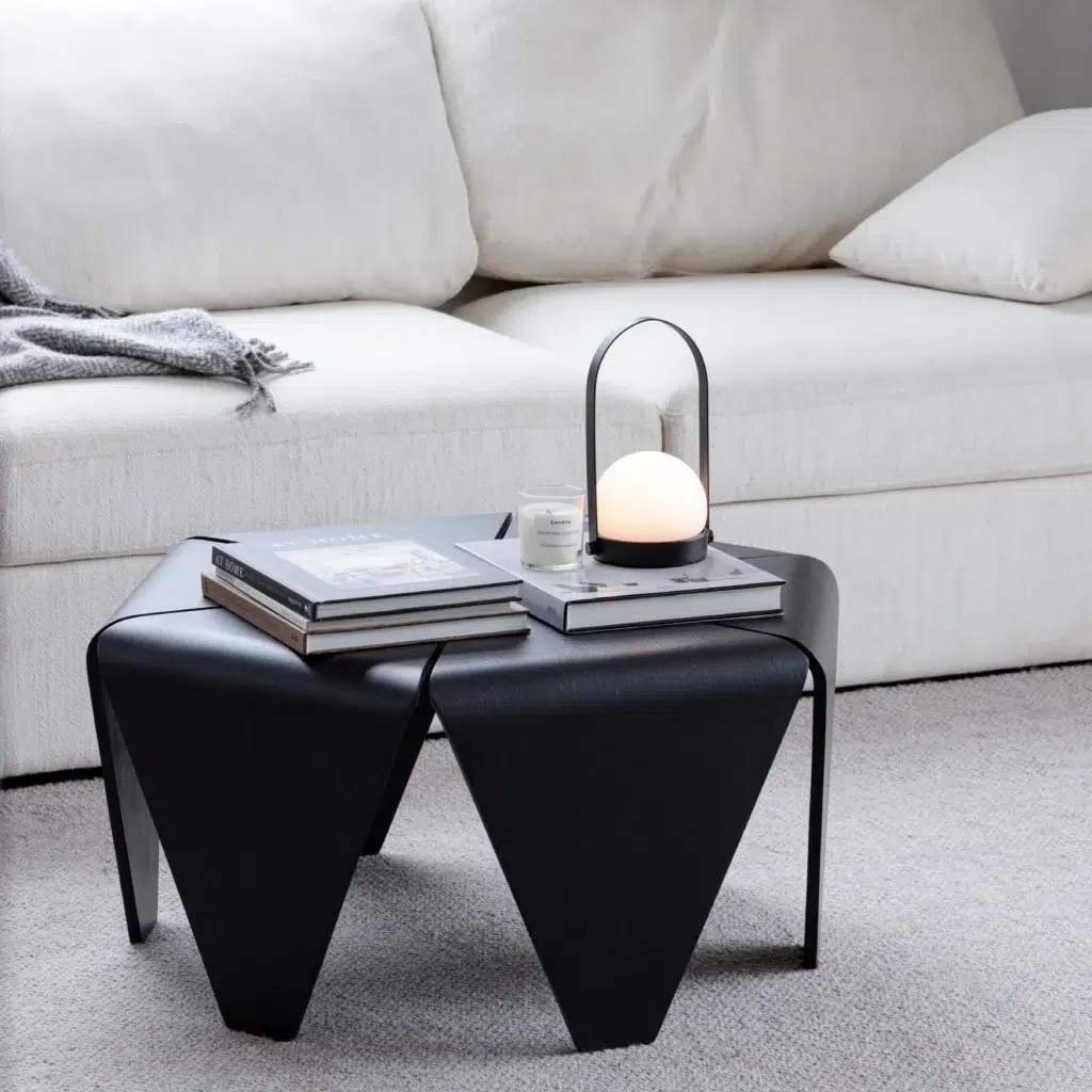 A modern black coffee table with books, a candle, and a round lamp sits on a light carpet in front of a white sofa with a grey throw blanket and cushions.