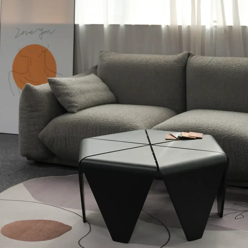 A modern living room with a grey textured sofa, a black geometric coffee table, and abstract artwork propped against the wall. Soft light filters through white curtains in the background.