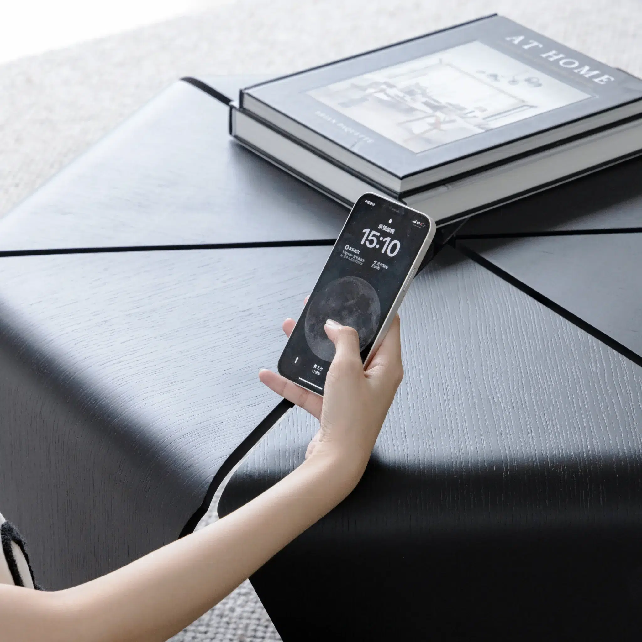 A hand holding a smartphone with a moon-themed lock screen is resting on a modern black coffee table. A large book titled AT HOME sits on the table in the background.