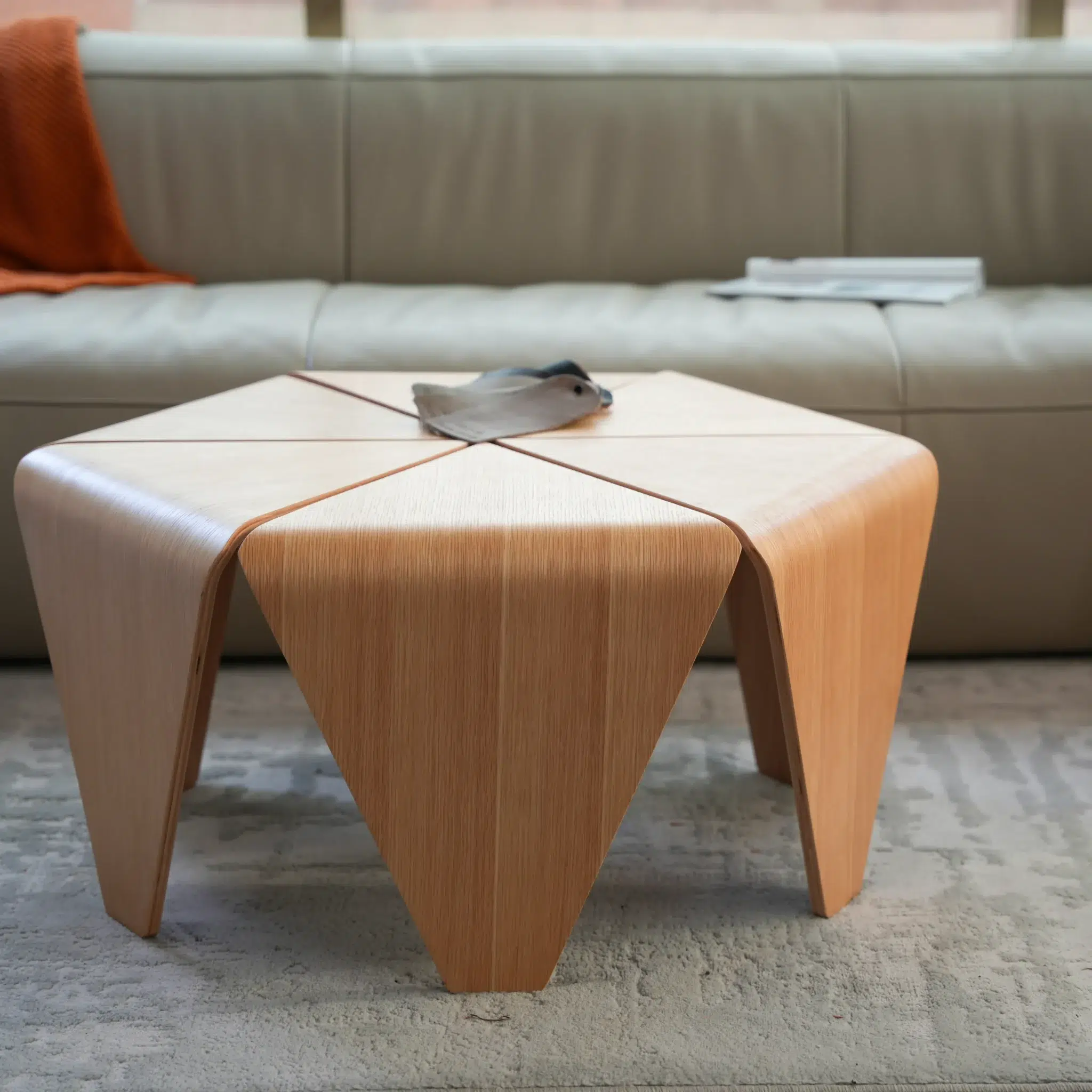 A modern, geometric wooden coffee table with triangular legs sits on a textured rug in front of a light grey sofa. An orange throw and an open magazine are on the sofa, with a small tray centred on the table.