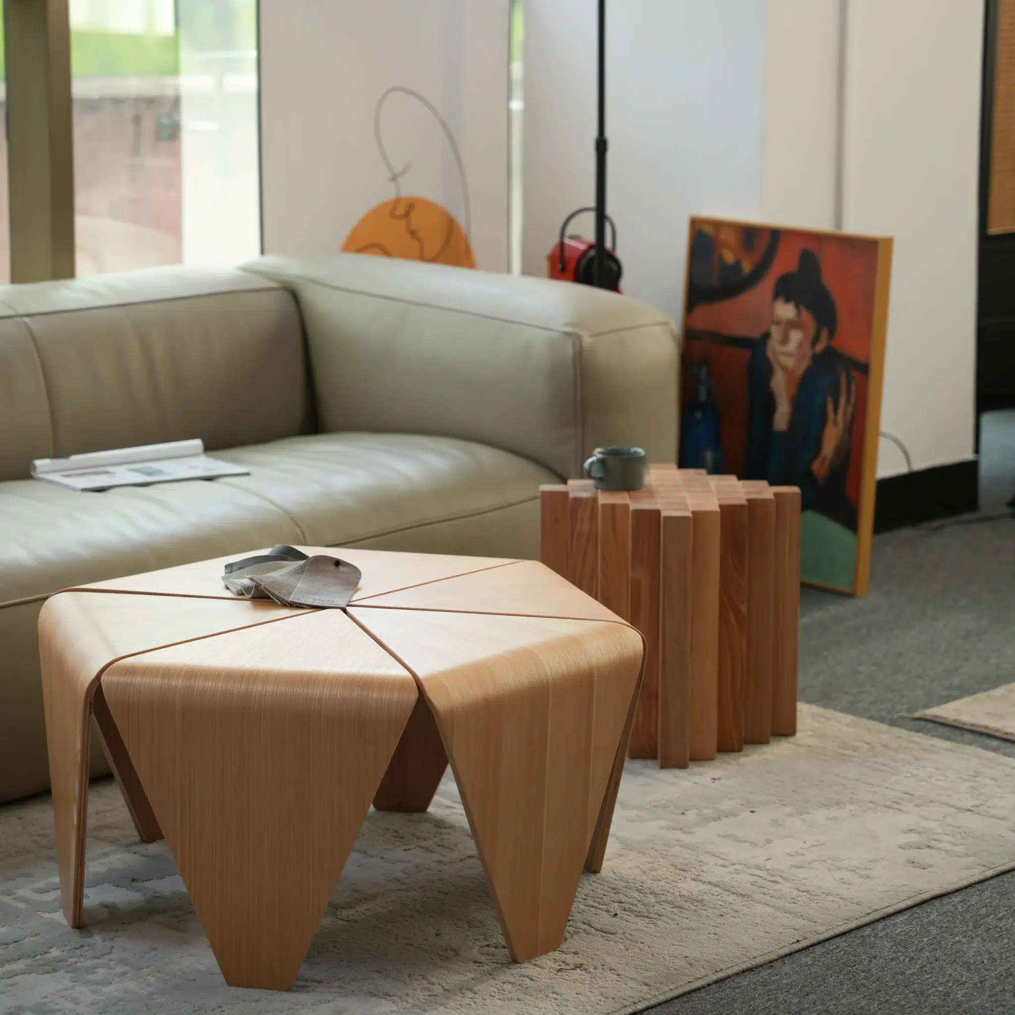 A modern living room with a light grey sofa, a unique wooden coffee table, a wooden side table, a cup, and a book. A painting and headphones rest near the sofa, and sunlight streams through a window.