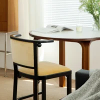 A wooden chair with a light yellow cushioned seat sits by a round wooden table. An open book, a candle, and a vase of green flowers rest on the table near a window with white blinds.