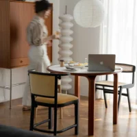 A blurred woman walks past a round dining table with food, a laptop, and two chairs in a sunlit, modern room with wooden floors and minimalist decor.