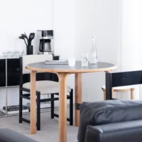 A round wooden dining table with two chairs, a glass bottle, drinking glasses, and a notebook sits in a bright, modern room. A coffee maker, utensils, and shelves with cups and books are in the background.