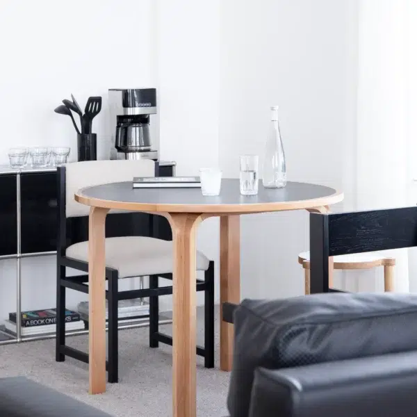 A round wooden dining table with two chairs, a glass bottle, drinking glasses, and a notebook sits in a bright, modern room. A coffee maker, utensils, and shelves with cups and books are in the background.