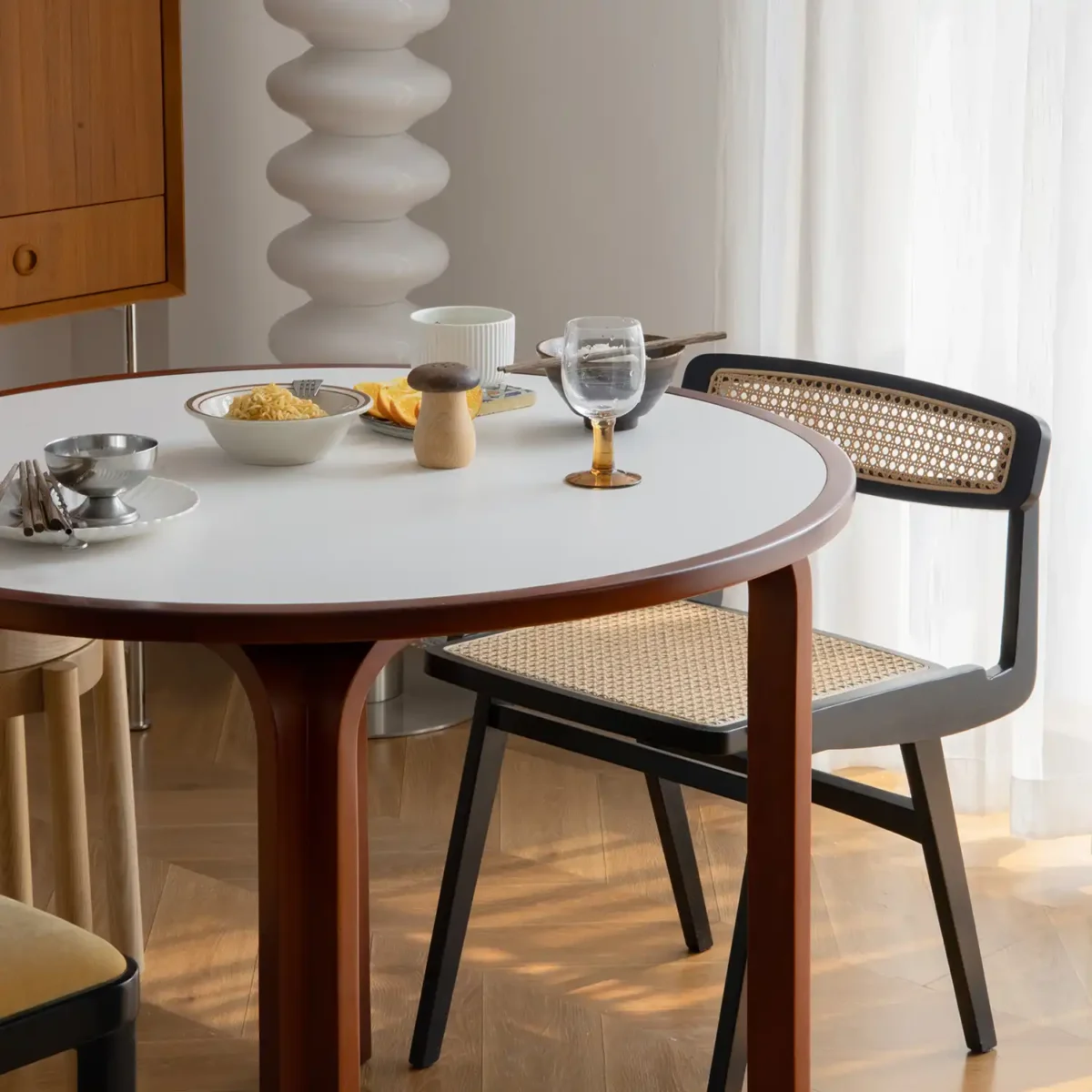 Loulan Round Dining Table_Front View_Detail A round dining table set with bowls, cups, and glasses beside a woven-back chair in a sunlit room with wooden floors and sheer white curtains.