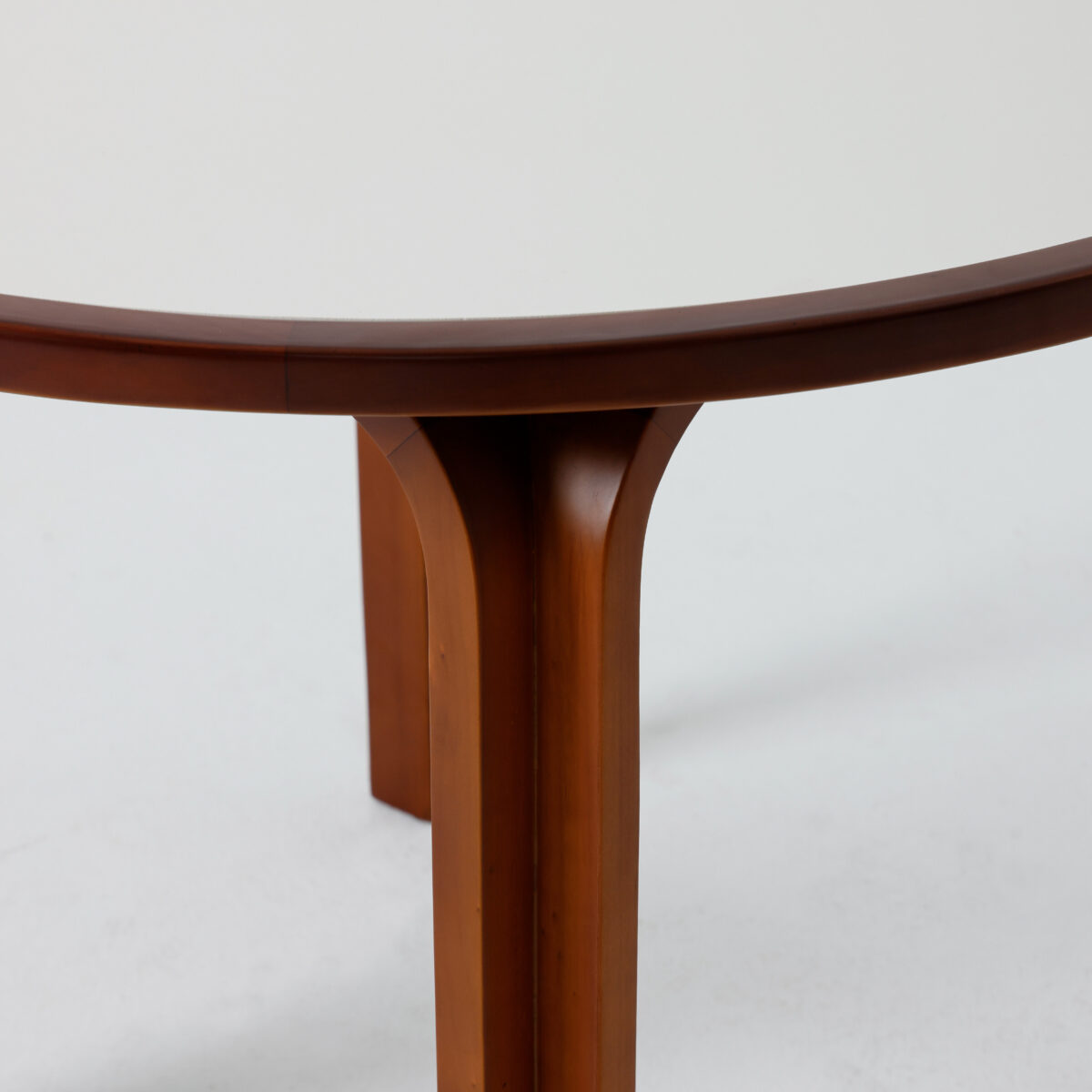 Loulan Round Table_Dark Brown_B3 Close-up of a round table with a white tabletop and wooden edge, showing two sturdy wooden legs joined at the centre and part of the curved underside. The background is plain and light-coloured.