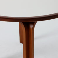 Close-up of a round table with a white tabletop and wooden edge, showing two sturdy wooden legs joined at the centre and part of the curved underside. The background is plain and light-coloured.