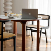 A round wooden table with a laptop, bowls of snacks, and cutlery on top, surrounded by two chairs on a wooden floor, with sunlight streaming in through sheer white curtains.