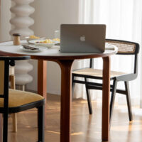 A round wooden table with a laptop, bowls of snacks, and cutlery on top, surrounded by two chairs on a wooden floor, with sunlight streaming in through sheer white curtains.