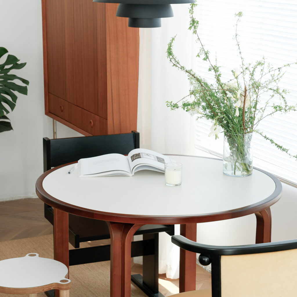 A round white table with wooden legs holds an open book, a lit candle, and a glass vase of green and white flowers. Black and cream chairs surround the table in a bright, modern room with natural light.